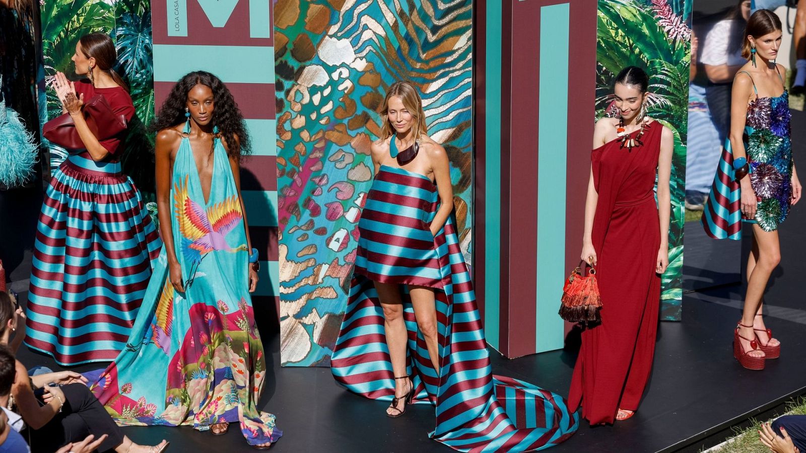 Desfile de moda de Lola Casademunt con modelos luciendo vestidos y faldas con estampados tropicales y rayas granates y turquesas. El fondo de la pasarela muestra el nombre de la marca.