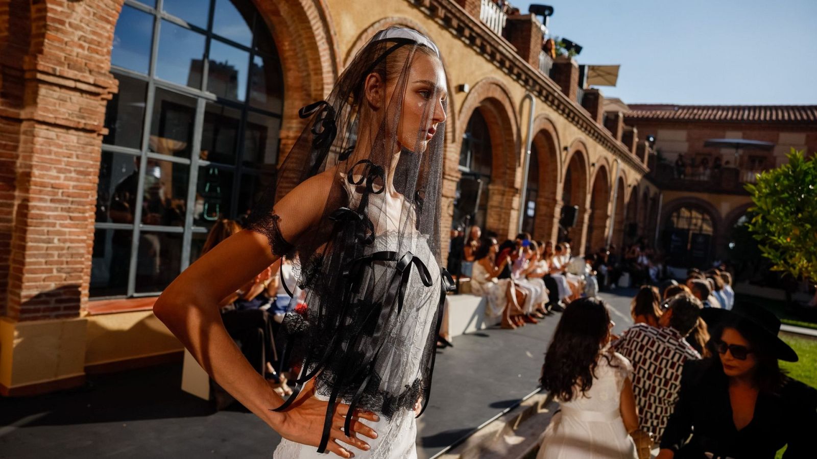 Modelo con velo y vestido Yolancris en desfile al aire libre, arquitectura de ladrillo visto y audiencia al fondo.