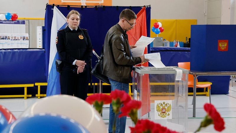 Hombre votando en centro electoral ruso. Urna con escudo de Rusia. Mujer con uniforme oficial y bandera rusa al fondo. Decoración con globos.