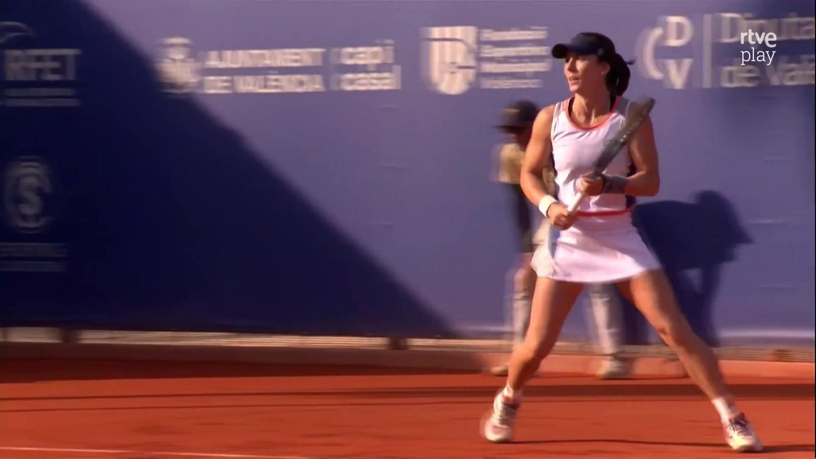 Tenista en cancha de tierra batida, con gorra azul y vestido blanco, lista para jugar. Fondo con "AJUNTAMENT DE VALÈNCIA" y "rtve play".
