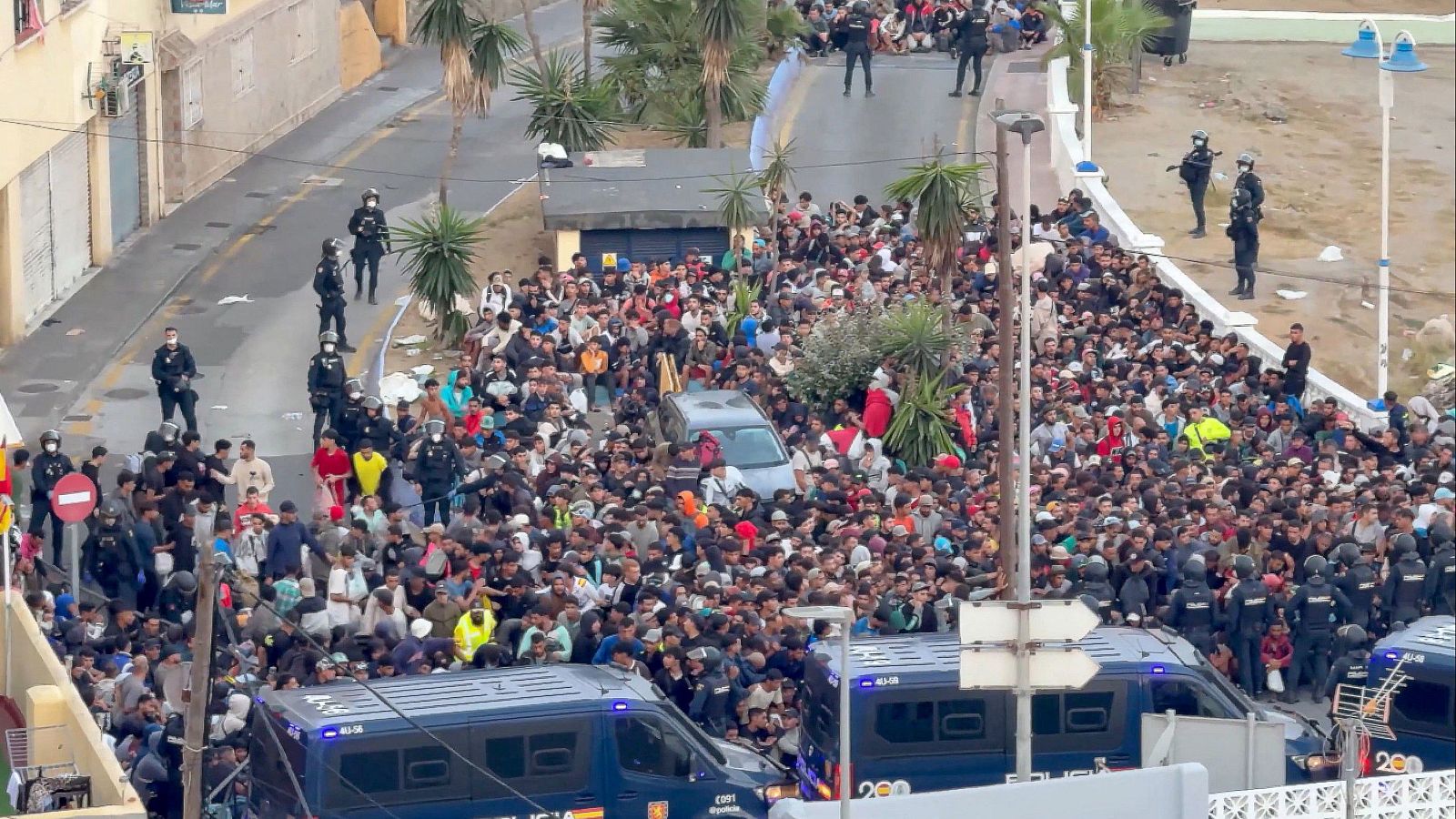 Miles de personas, en su mayoría hombres jóvenes, abarrotan una calle urbana, flanqueados por agentes de la Policía Nacional y furgones policiales. Se observa un coche civil rodeado por la multitud.