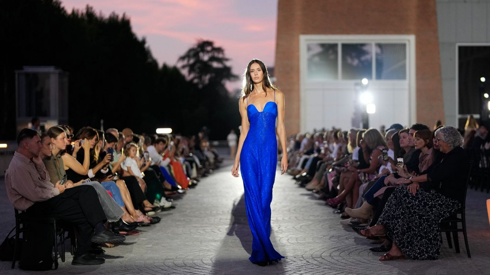 Desfile de moda al atardecer: modelo con vestido azul eléctrico brillante en pasarela, audiencia atenta a ambos lados. Cielo crepuscular y edificios modernos al fondo.