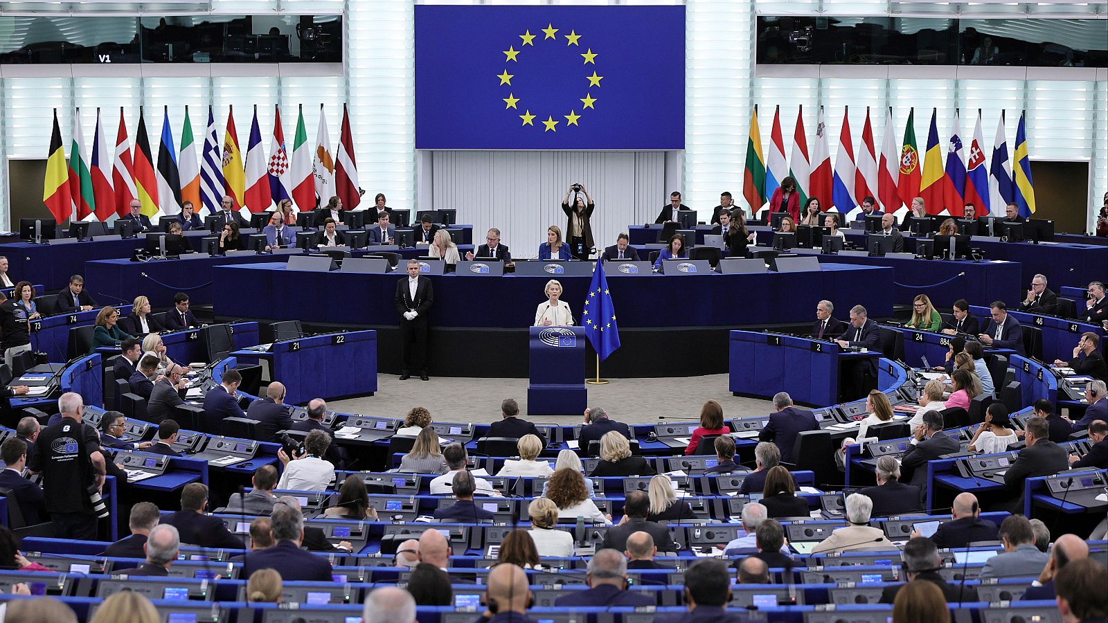 Ursula von der Leyen habla en el Parlamento Europeo, rodeada de banderas de la UE y de estados miembros, ante una audiencia de eurodiputados.