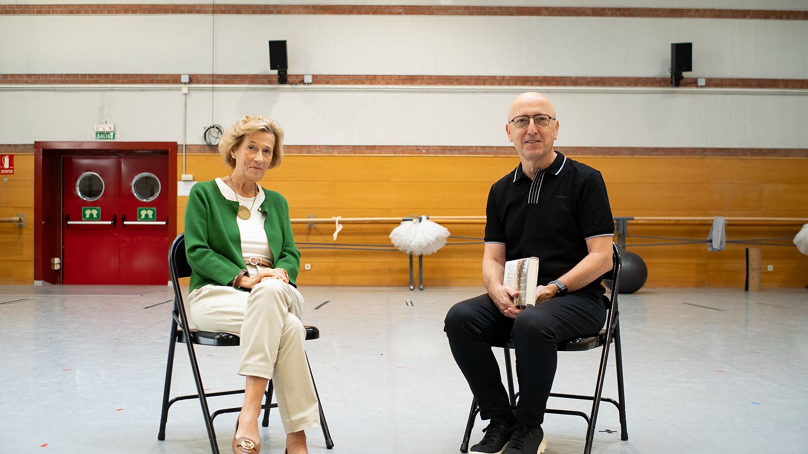 Julia Navarro, con chaqueta verde y collar dorado, y Óscar López, con polo negro y gafas, posan en un estudio con elementos de ballet y salida de emergencia al fondo.