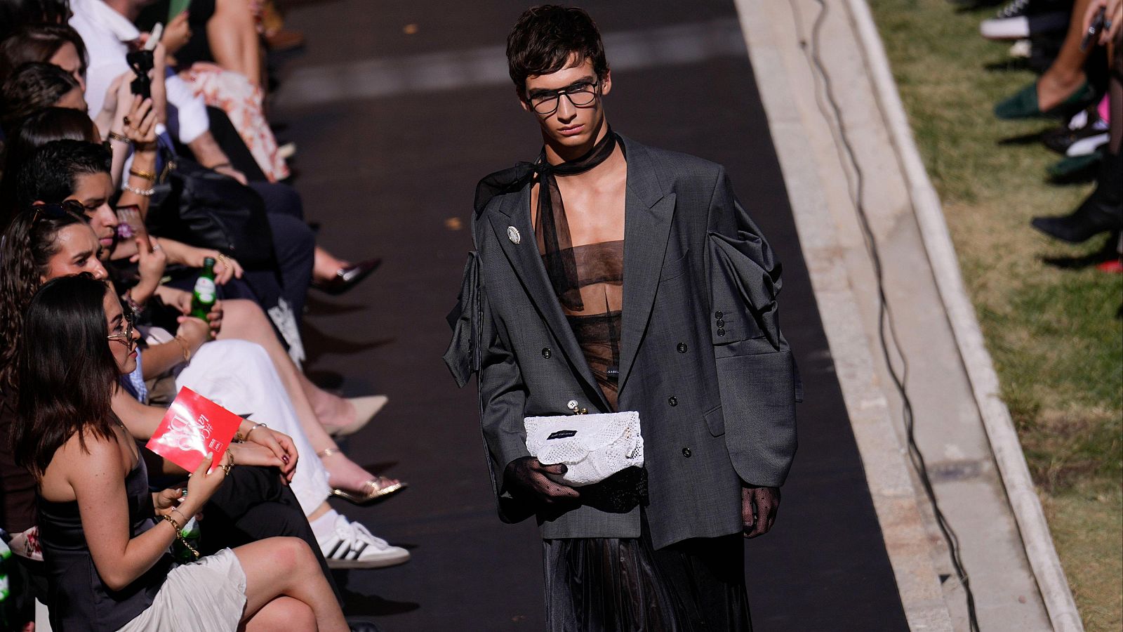 Modelo masculino con blazer gris deconstruido, top y guantes transparentes, camina por pasarela. Espectadores con móviles observan. Césped al lado.