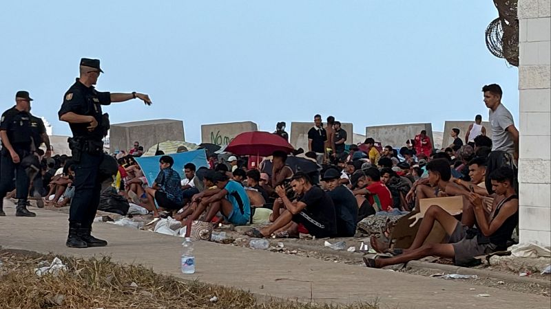Agentes uniformados supervisan a un grupo de hombres jóvenes sentados en el suelo, protegidos por refugios improvisados, con barreras al fondo.