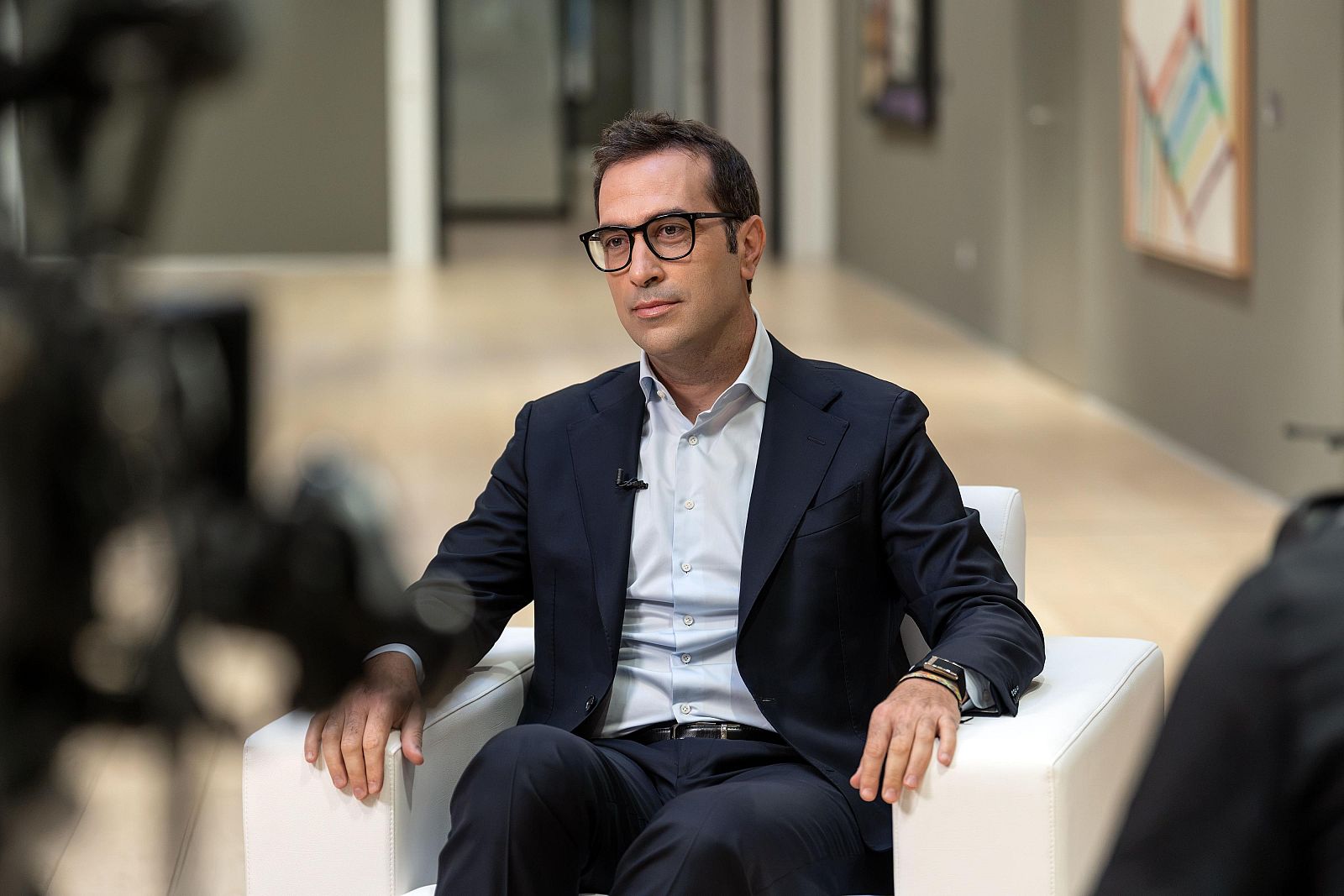 Carlos Cuerpo, con gafas y traje azul, sentado en sillón blanco, con micrófono de solapa. Fondo con arte abstracto.