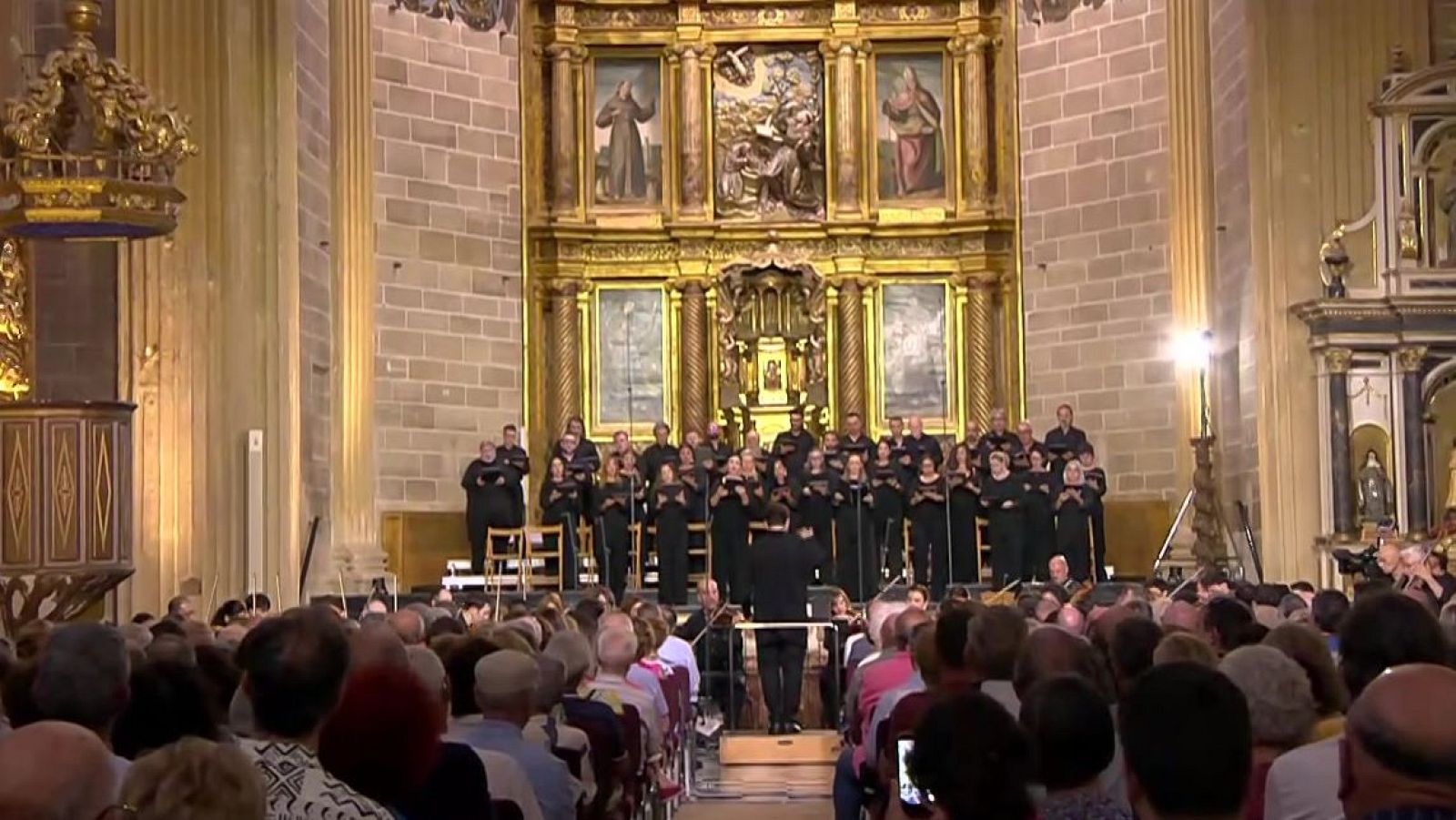 Concierto de música clásica en una iglesia, con coro, orquesta y director. El público asiste en la nave central del templo.