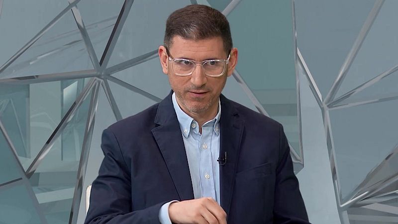 Primer plano de José María Medina, periodista y presentador de TVE, con gafas, barba y traje azul. Fondo abstracto de paneles geométricos.