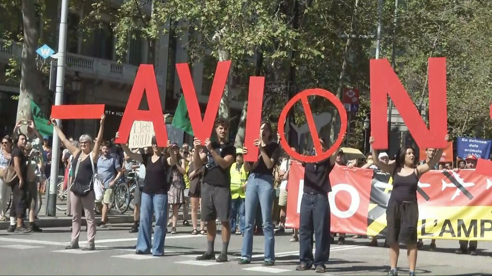 Manifestación "NO AVIÓN" con lema "En defensa dels animals" y símbolo de avión tachado. Calle urbana con árboles y edificios. Participantes con ropa casual. Cataluña, España.