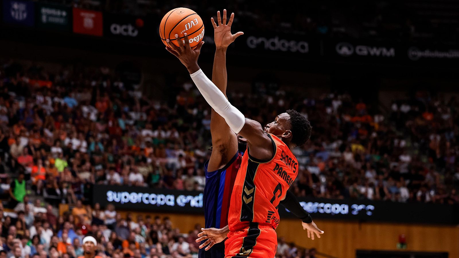 Jugador con camiseta naranja "SHORTS PAMER" y dorsal "0" lanza a canasta "EVO" en la "SUPERCOPA ENDESA", con público y banners "Endesa", "act", "GWM" de fondo.