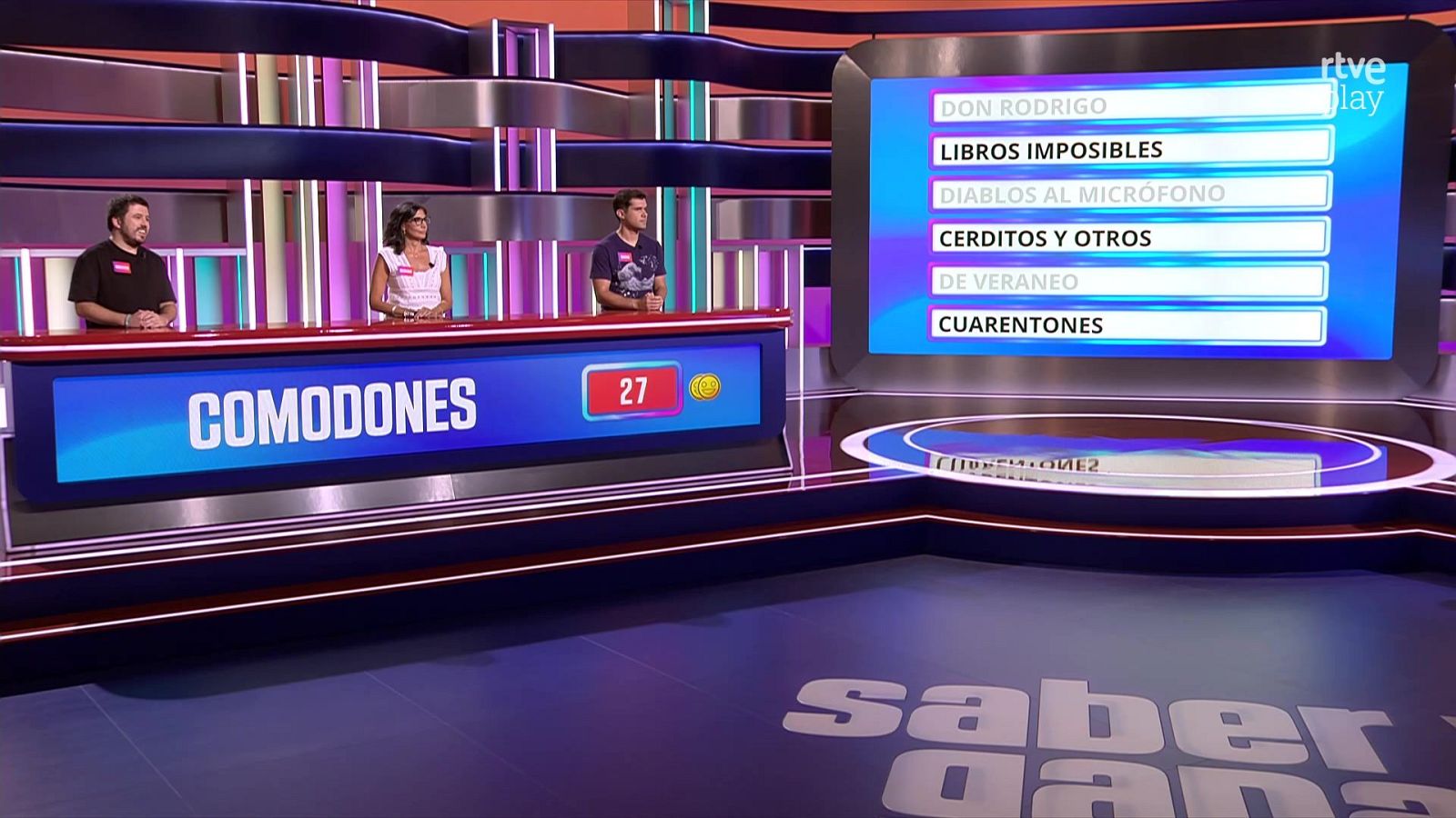 Tres concursantes en un plató de "Saber y Ganar" con el mostrador "COMODONES" y la pantalla de categorías "DON RODRIGO", "LIBROS IMPOSIBLES", "DIABLOS AL MICRÓFONO", "CERDITOS Y OTROS", "DE VERANEO", "CUARENTONES".