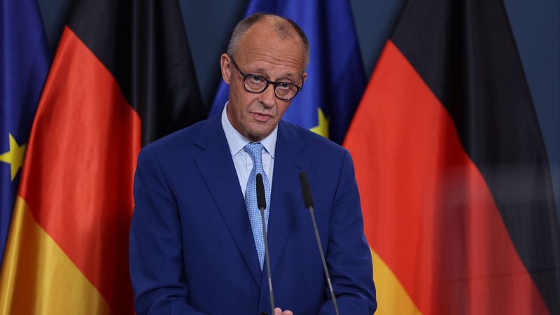 Primer plano de Friedrich Merz, líder de la CDU, con traje azul y gafas, hablando detrás de un atril con las banderas de la UE y Alemania.