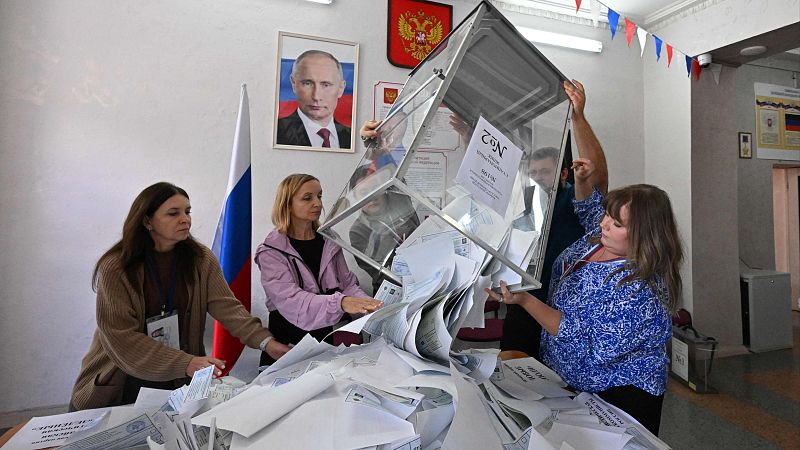 Mujeres rusas vacían una urna electoral "№2" en un colegio electoral. El fondo exhibe símbolos nacionales y un retrato de Vladimir Putin.