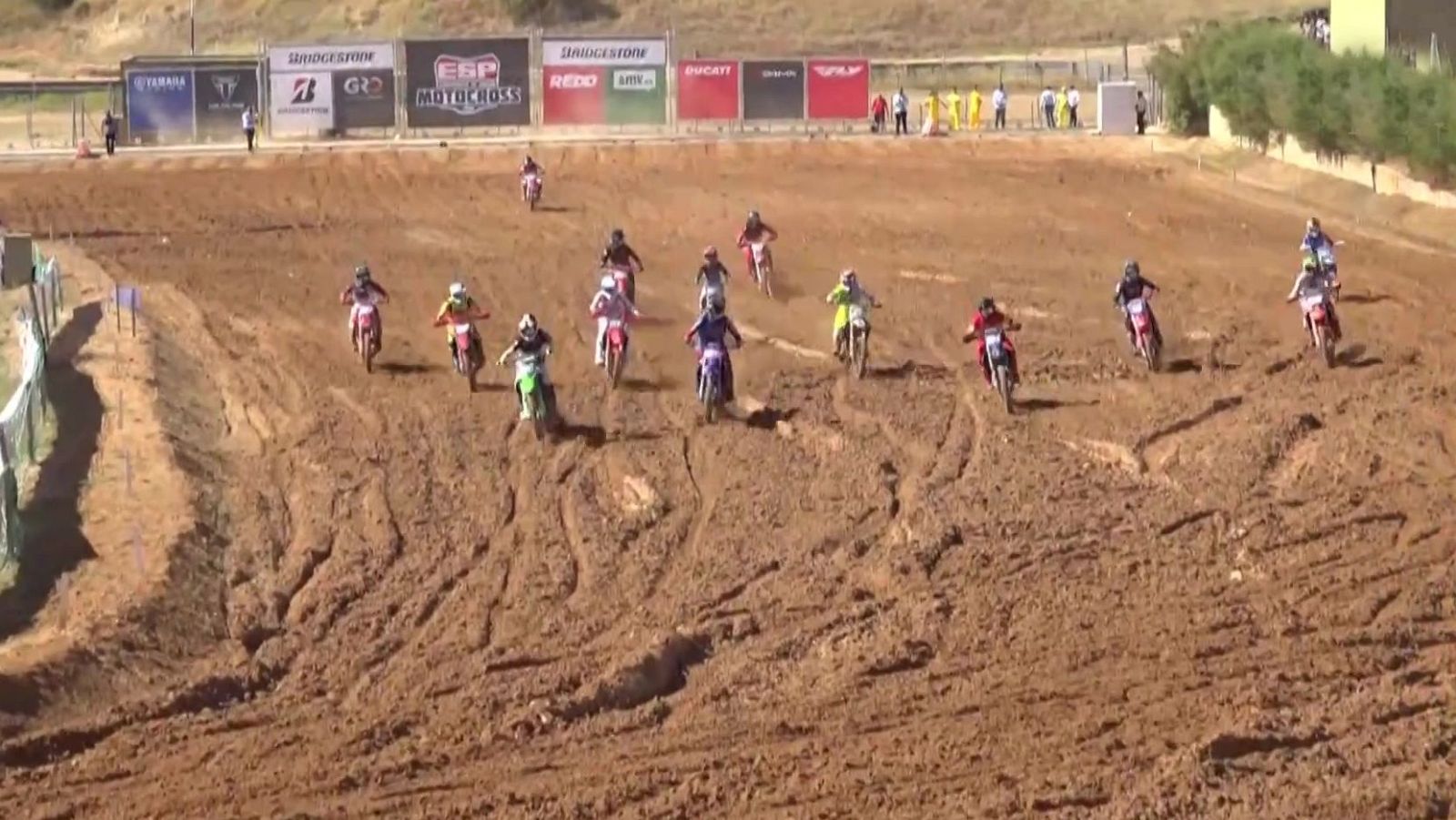 Carrera de motocross con múltiples pilotos en un circuito de tierra. Se aprecian paneles publicitarios de marcas como Yamaha, Bridgestone y Ducati al fondo.
