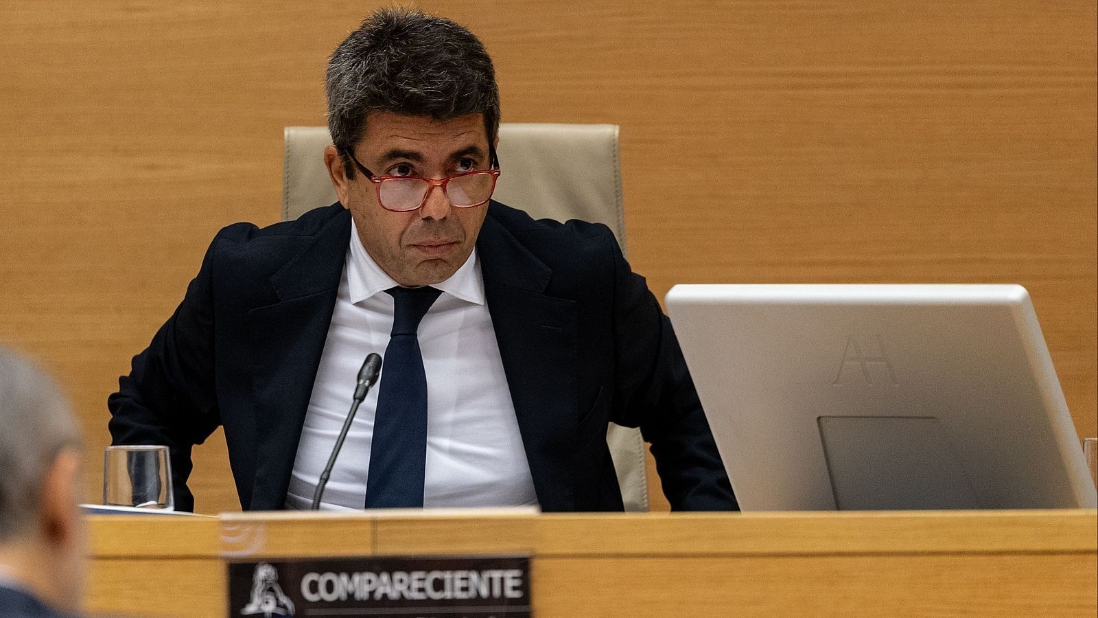 Carlos Mazón, compareciente en el Congreso, con gafas rojas y traje oscuro, sentado ante un micrófono y un dispositivo "AH".
