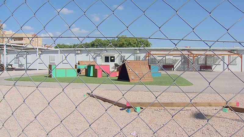 Vista exterior de un patio escolar con barracones y juegos infantiles, capturada a través de una valla metálica. Césped artificial, bloques de colores y estructuras de madera componen la zona de recreo.