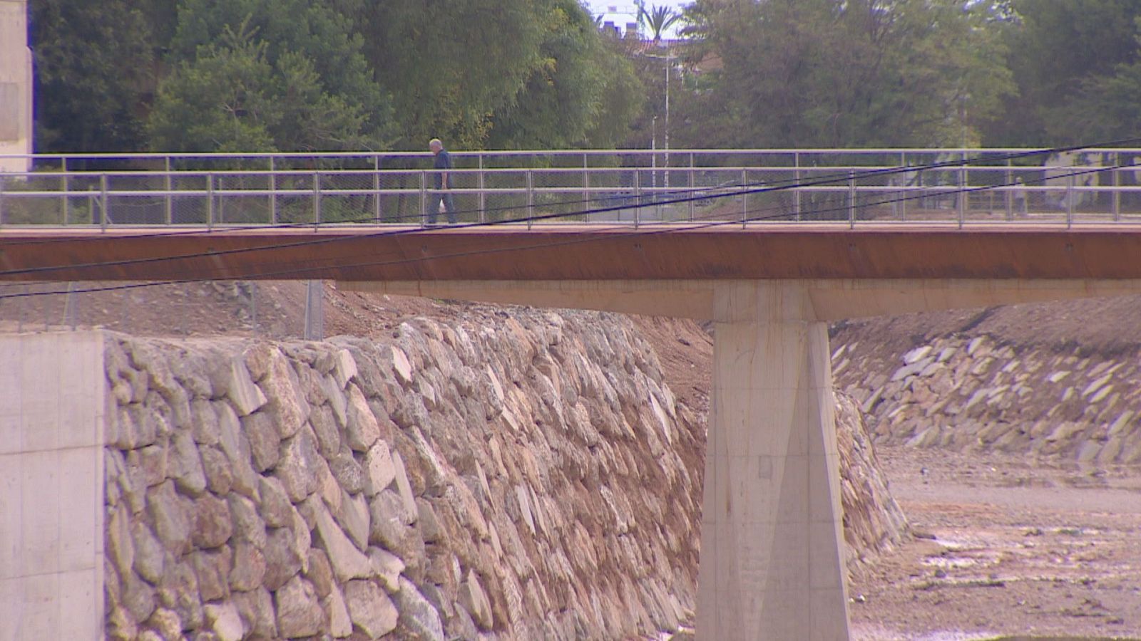 Puente moderno de acero corten y hormigón cruza un cauce seco con muros de piedra. Una persona camina sobre él. Vegetación y edificaciones al fondo.