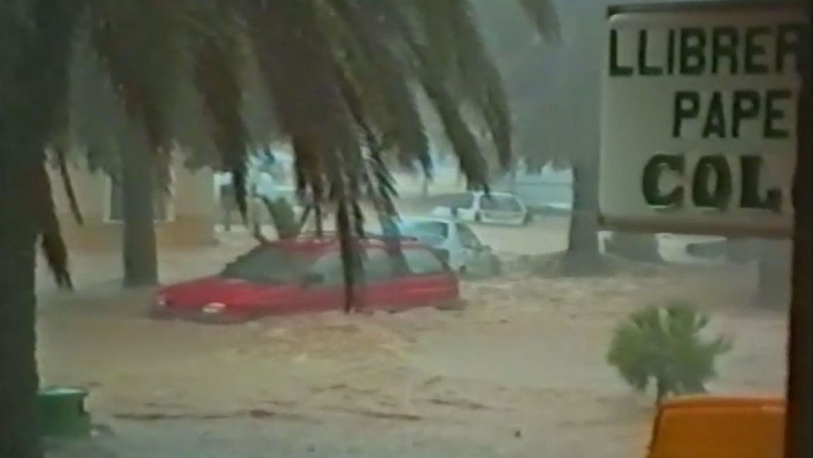 Calle inundada en la Comunidad Valenciana con vehículos sumergidos, palmeras y un cartel que indica 'LLIBRER', 'PAPE', 'COL'.