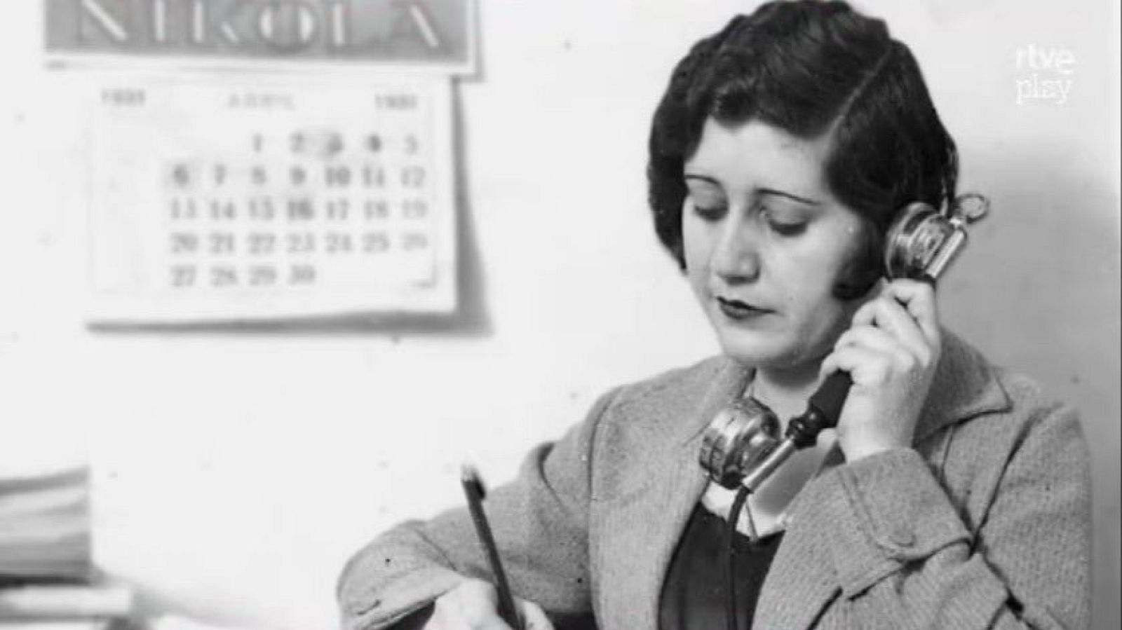 Josefina Carabias, periodista, en un primer plano de medio cuerpo, con un teléfono antiguo y bolígrafo, tomando notas. Calendario de abril de 1931 al fondo. Marca de agua 'rtve play'.