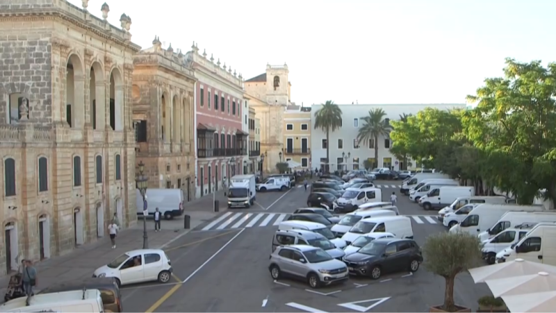 Vista aérea de la Plaça del Born, Ciutadella de Menorca. Edificio histórico, aparcamiento con furgonetas y coches, y torre de campanario. Actividad urbana con personas y vehículos.
