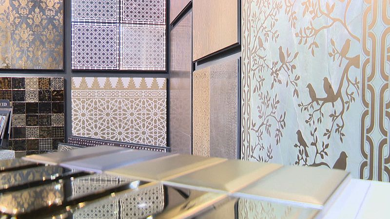 Exposición de azulejos y revestimientos con diseños geométricos, damascos y motivos de pájaros en tonos dorados y metálicos. Muestras reflectantes en primer plano.
