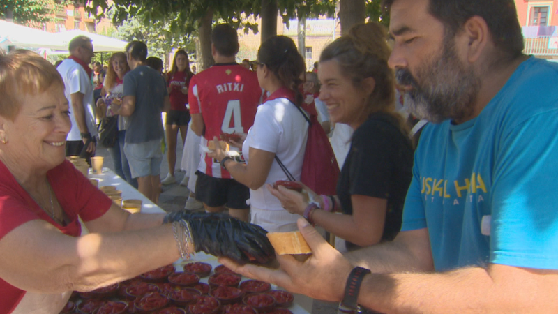 Degustación popular de pimientos del piquillo con personas sirviendo y recibiendo raciones. Se observa una camiseta del Athletic Club y un ambiente festivo.