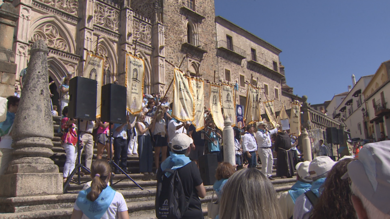 Multitud reunida en la escalinata del Real Monasterio de Santa María de Guadalupe, con personas en hábitos y vestimentas blancas, portando pañuelos azules y estandartes. Se aprecian altavoces y un pilar ornamentado.