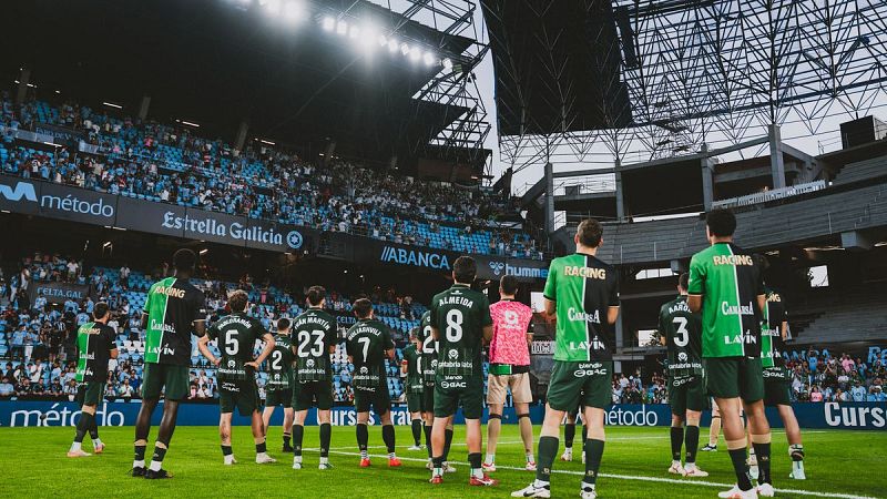 Jugadores de fútbol con uniformes "RACING" en un estadio, con nombres como "ALMEIDA" y "JUAN MARTIN" visibles. Foco en el césped, gradas y vallas publicitarias.