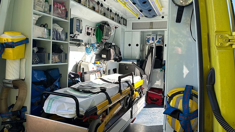 Interior de ambulancia equipada: camilla con logo "SESCAM", estanterías de suministros médicos, casco amarillo, y dispositivos de soporte vital. Espacio diseñado para atención de emergencias.