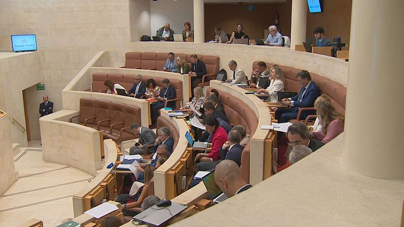 Vista del hemiciclo del Parlamento de Cantabria con asistentes en sesión, equipados con ordenadores y documentos. Se observa una escalera y una señal de salida de emergencia.