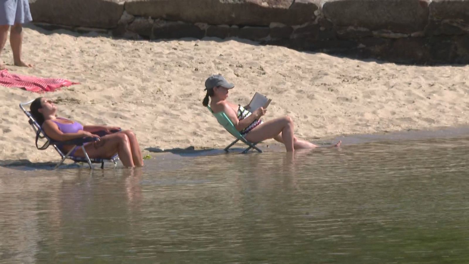 Mujeres en sillas de playa en la orilla, una leyendo y otra descansando, en un día soleado.
