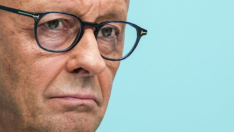 Friedrich Merz, con gafas oscuras y expresión seria, en un primer plano con fondo azul claro. Se aprecian arrugas y un logo en la patilla de las gafas.