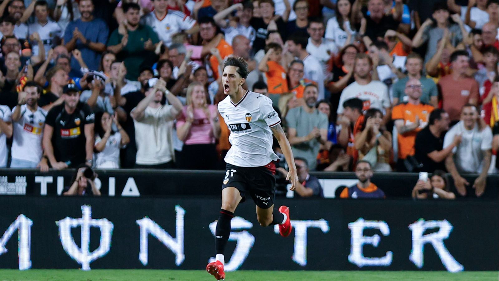 Fran Pérez (Valencia CF, dorsal 17) celebra un gol con euforia. Viste uniforme blanco y negro, medias rojas. Multitud anima en gradas. Valla publicitaria "MONSTER" al fondo.
