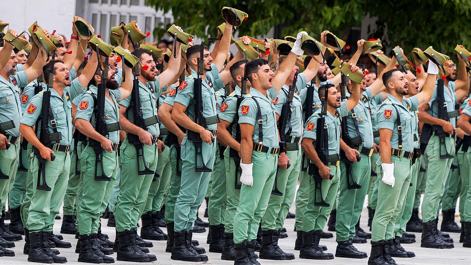 Soldados de la Legión Española en Melilla, uniformados de verde, con gorros cuarteleros y rifles, cantan o gritan con fervor en un acto militar.