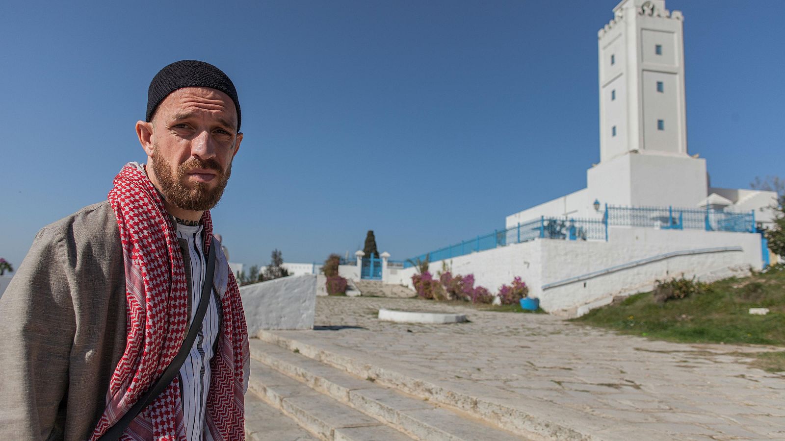 Activista Fergie Chambers frente a una mezquita blanca con minarete en Túnez, bajo un cielo azul.