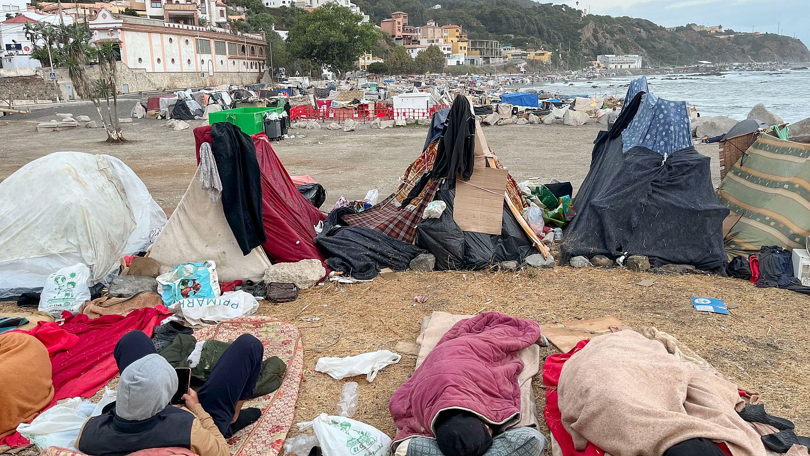 Campamento costero con personas descansando y refugios improvisados. Al fondo, el mar y edificaciones en una colina.