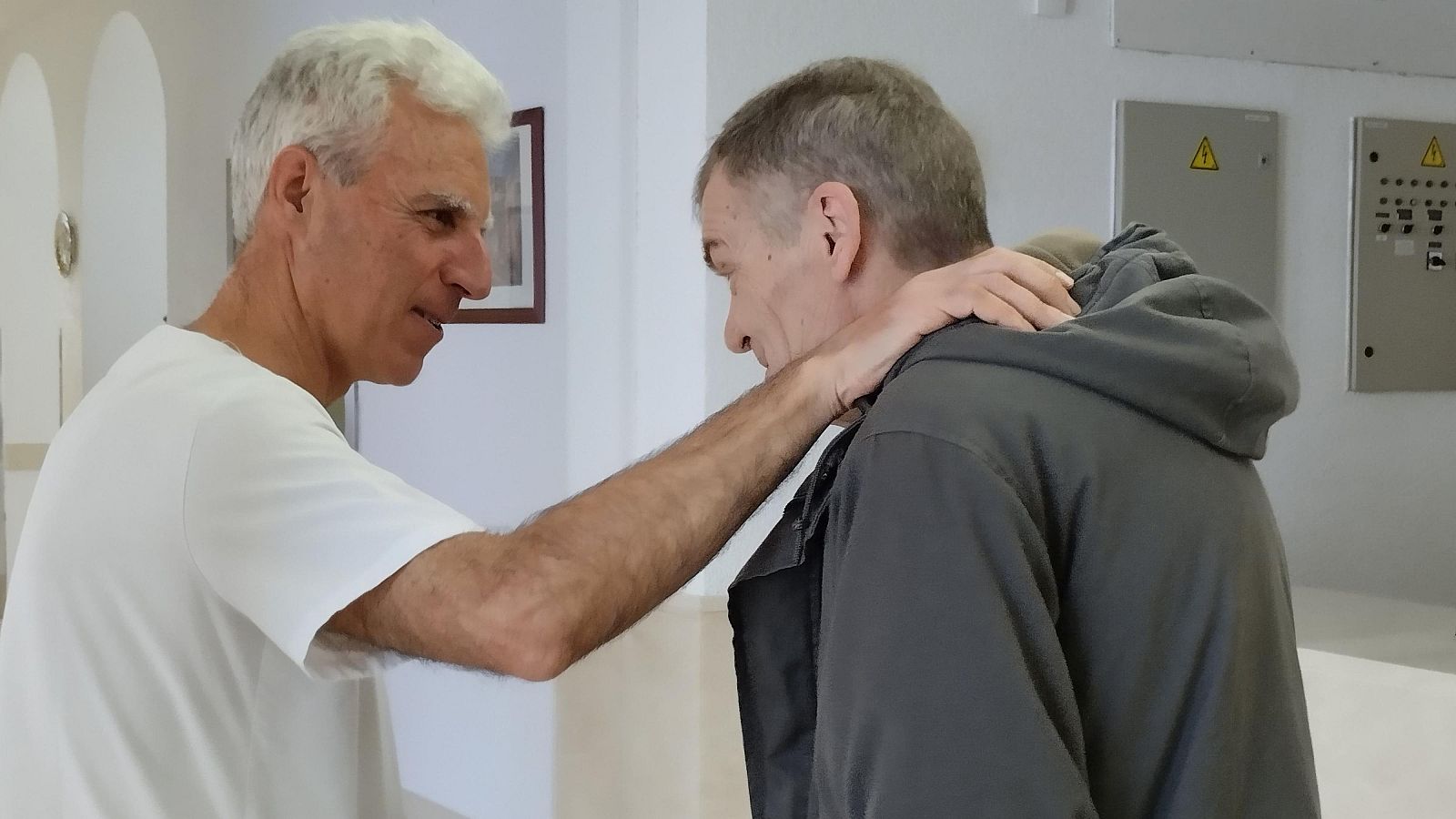 Primer plano de Miguel Bosé y otro hombre en interior. Bosé, de perfil, con camiseta blanca, apoya su mano en el hombro del otro hombre, que viste sudadera gris.