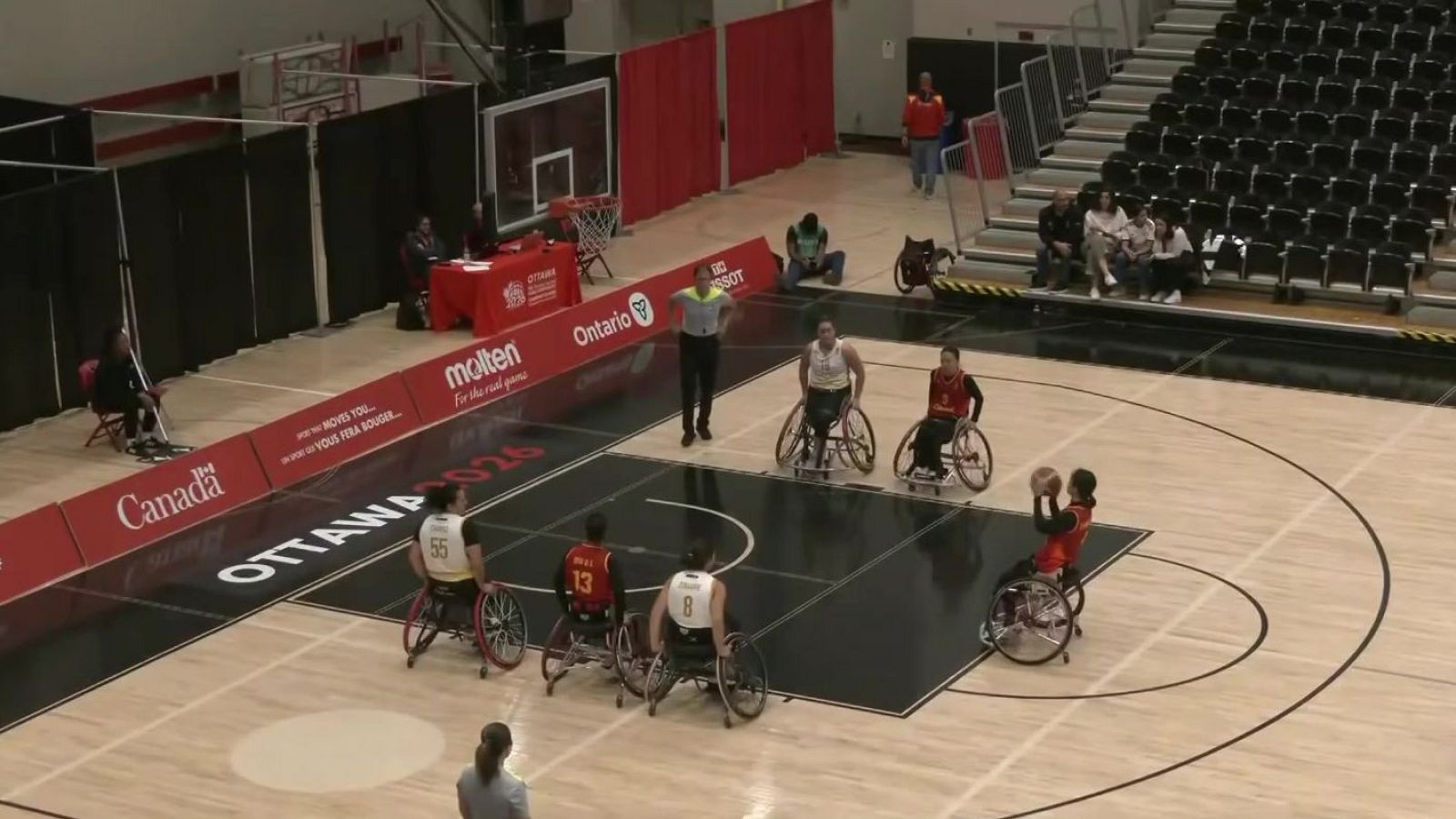 Vista aérea de un partido de baloncesto en silla de ruedas con árbitro y jugadores en acción. Se aprecian vallas publicitarias con "Canada" y "OTTAWA 2026".