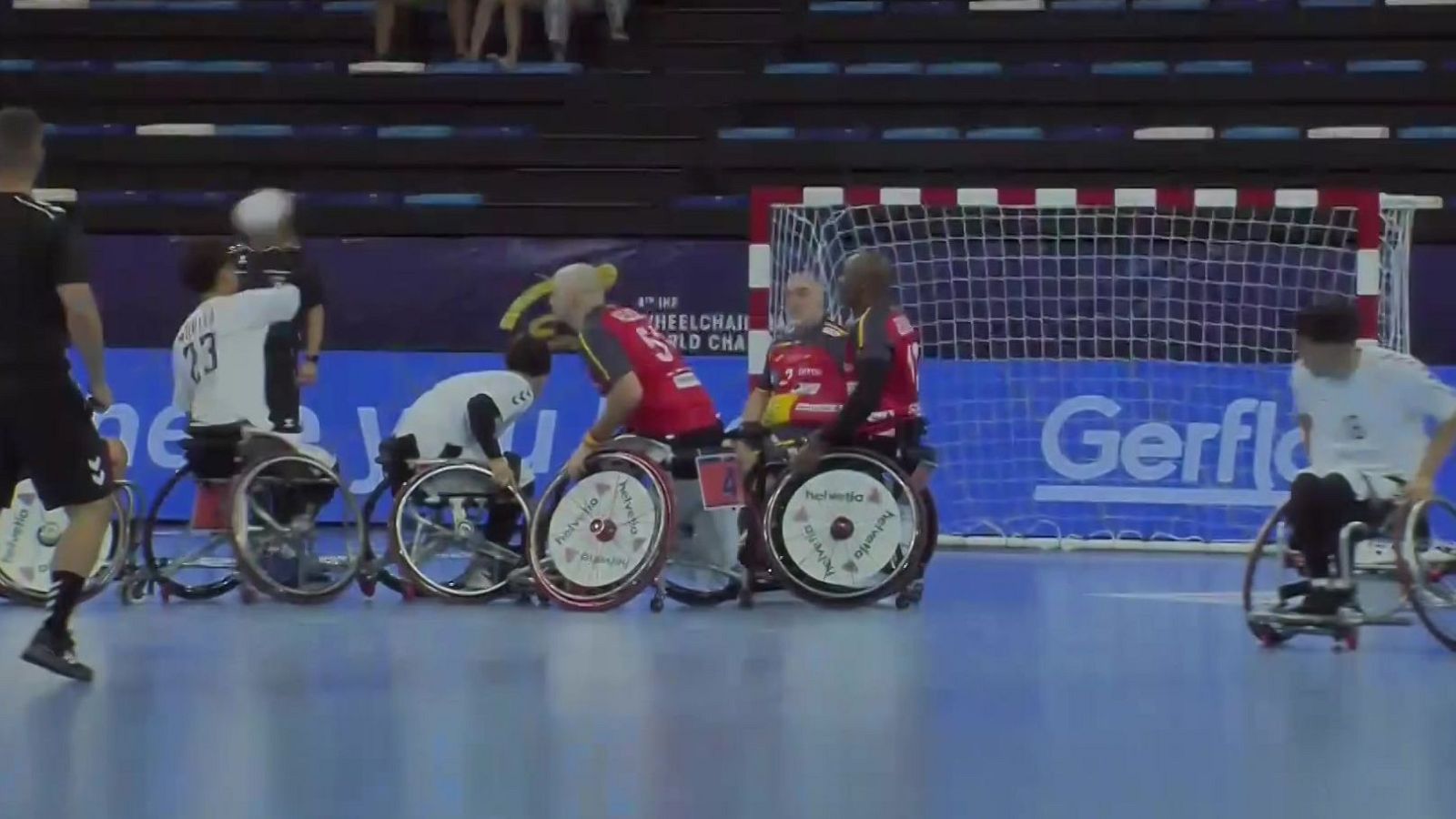 Jugadores de balonmano en silla de ruedas compiten en una cancha azul, vistiendo camisetas blancas y rojas. Se aprecian vallas publicitarias y la portería.