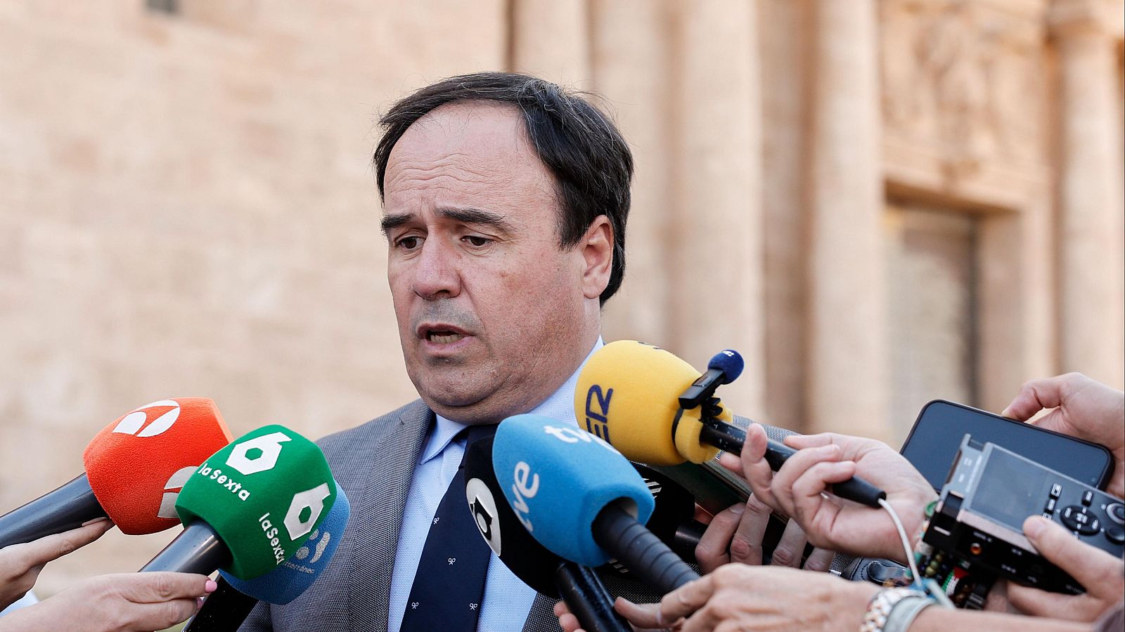 Carlos Mazón, Presidente de la Generalitat Valenciana, habla ante la prensa con micrófonos de laSexta, TVE y SER. Fondo de edificio institucional.