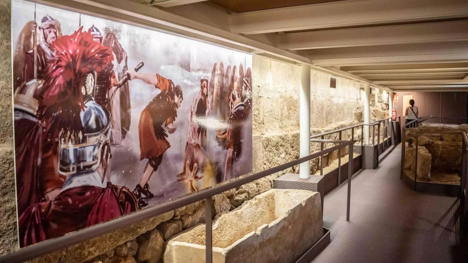 Interior de un sitio arqueológico con mural de soldados romanos, pasarela moderna y restos de piedra antiguos, incluyendo un posible sarcófago.