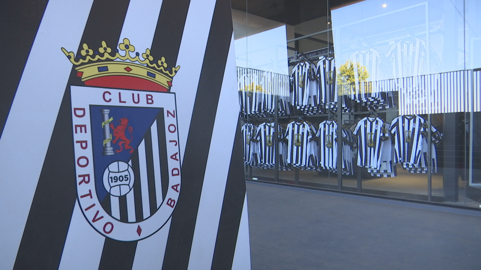 Escudo del Club Deportivo Badajoz con pilar, león rampante y balón de fútbol. Escaparate con camisetas a rayas blancas y negras.
