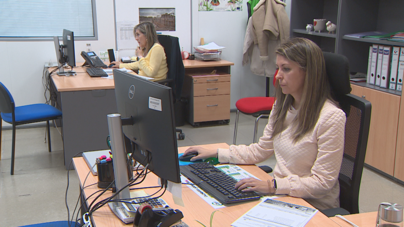 Dos mujeres trabajando en una oficina moderna. Una en primer plano usa ratón y teclado, la otra atiende una llamada telefónica. Ambas concentradas en sus monitores.