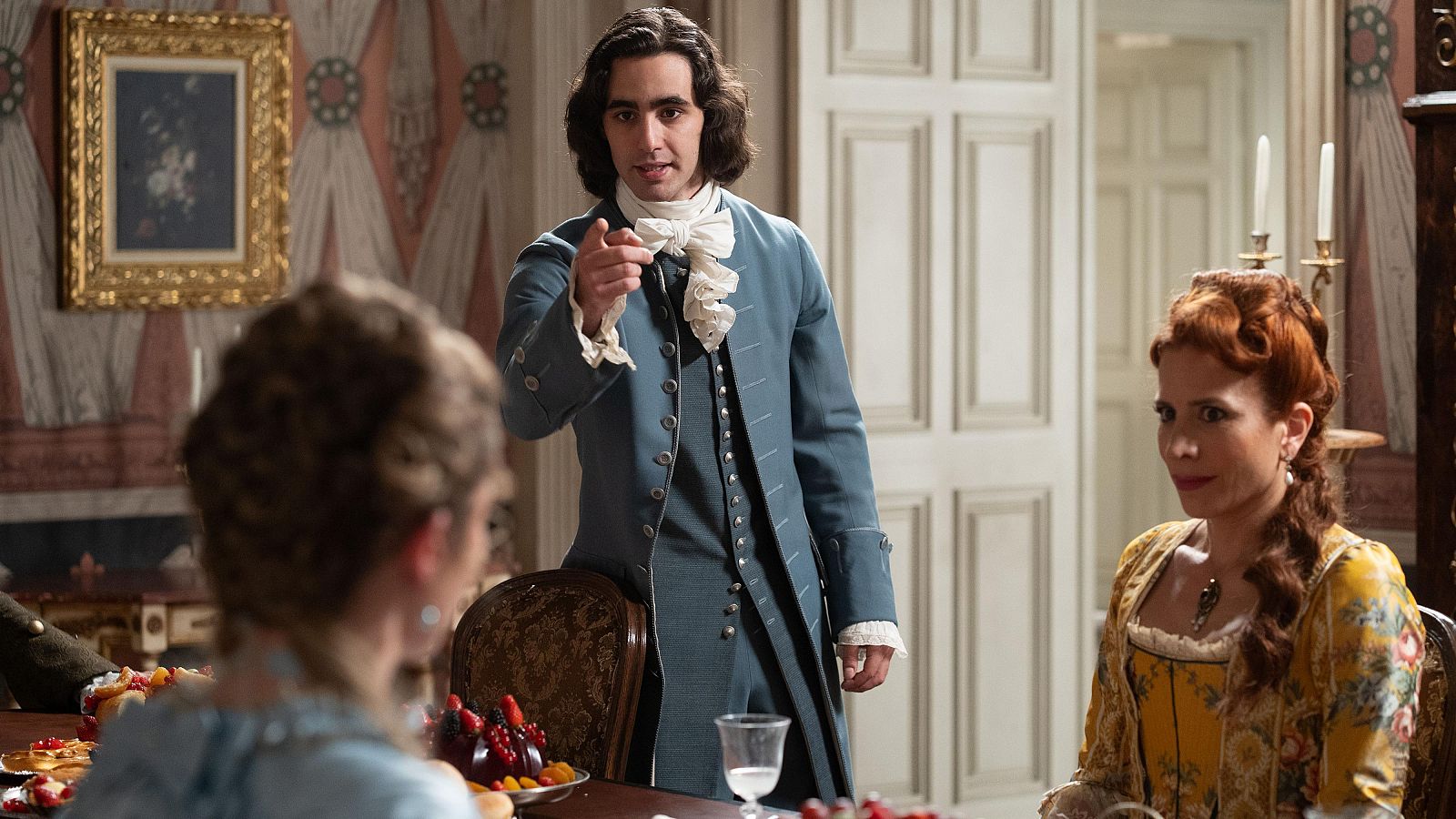 Alejo, con traje del siglo XVIII, señala con el dedo índice mientras una mujer pelirroja lo observa. La mesa está llena de postres y frutas. Escena de drama de época.