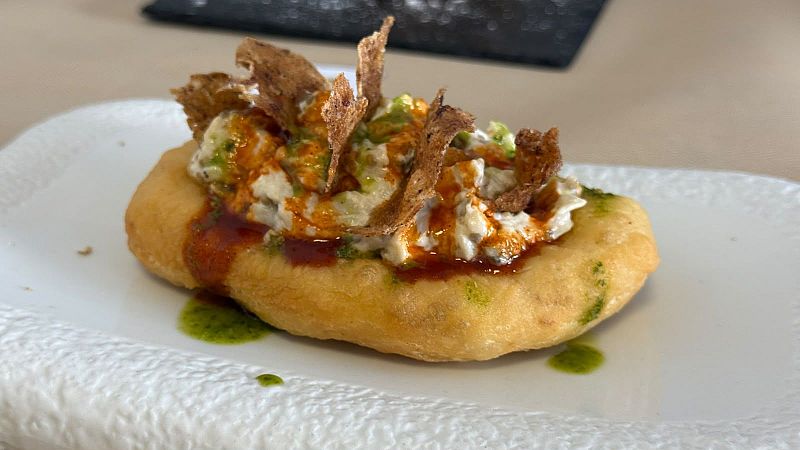 Coca ganadora del primer premio, con base dorada, topping cremoso blanco, salsa rojiza y verde, y crujientes trozos marrones, presentada en plato de cerámica blanca.