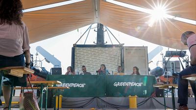 Evento de Greenpeace al aire libre bajo un toldo beige. Cinco ponentes en una mesa con mantel verde y logo de Greenpeace. Dos asistentes en primer plano. Entorno de muelle o barco.