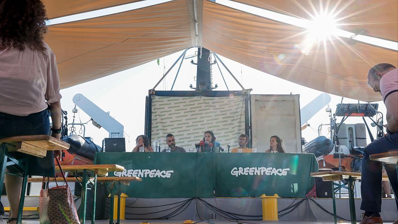 Evento de Greenpeace al aire libre bajo un toldo beige. Cinco ponentes en una mesa con mantel verde y logo de Greenpeace. Dos asistentes en primer plano. Entorno de muelle o barco.
