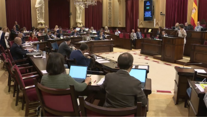 Sesión plenaria en el Parlamento Balear con diputados, una oradora en el atril y las banderas de España y Baleares. Ambiente institucional y tecnológico.
