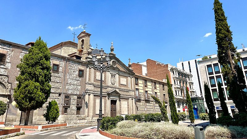 Vista del Monasterio de las Descalzas Reales en Madrid, con jardín, farol clásico, árboles y edificios históricos y modernos. Cielo azul y soleado.